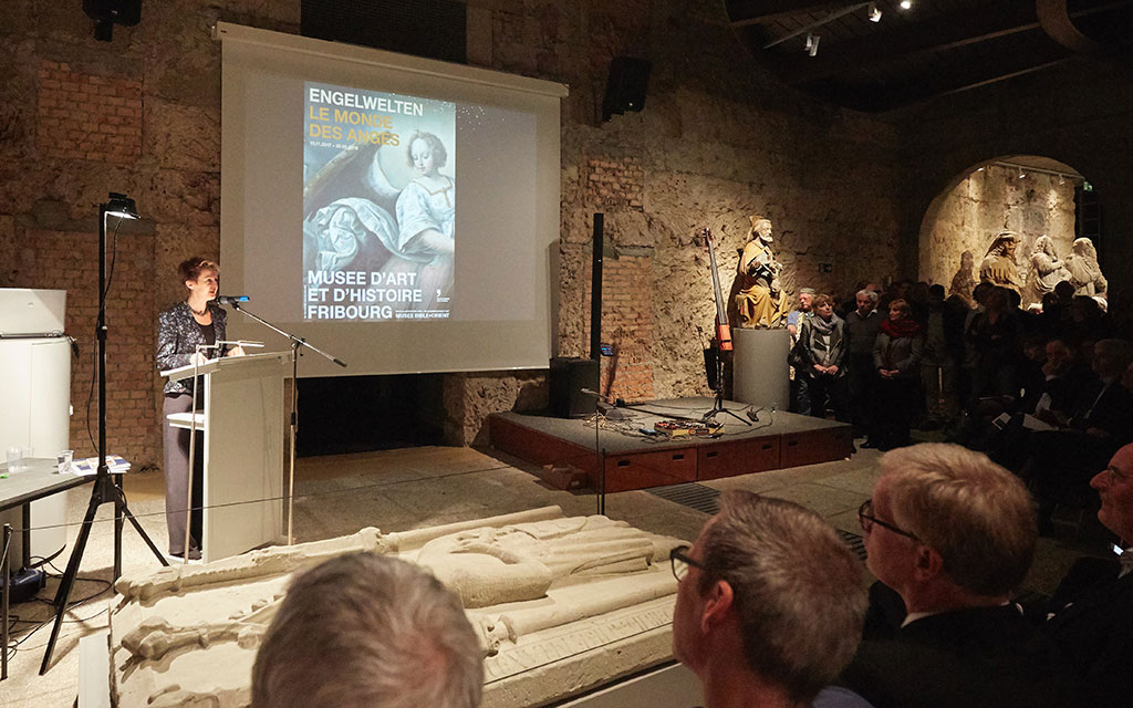 Bundesrätin Simonetta Sommaruga steht am Rednerpult und spricht in ein Mikrofon. Im Hintergrund ist das Plakat der Ausstellung "Engelwelten - Le monde des anges" des Kunstmuseums Freiburg zu sehen.